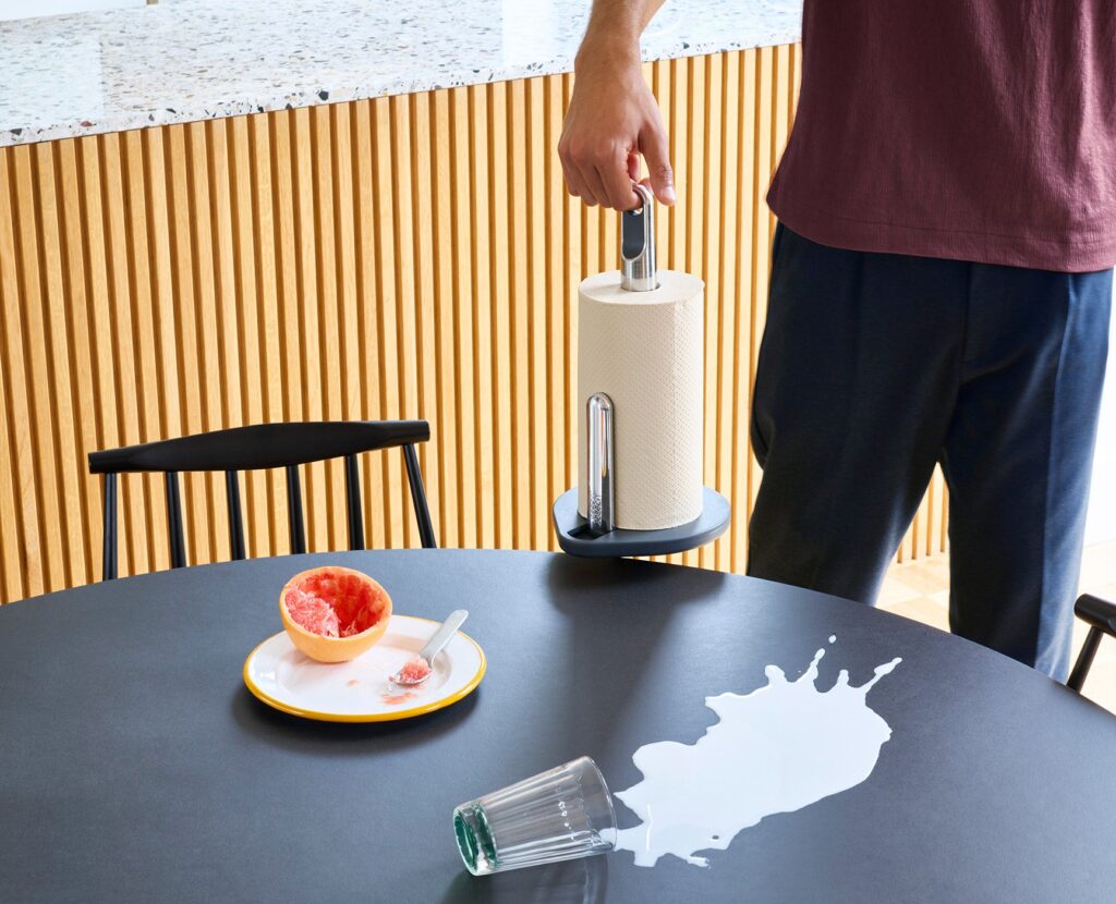 Joseph Joseph Hold Küchenrollenhalter wird an seiner Fingerschlaufe zu verschütteter Milch auf einem Tisch getragen