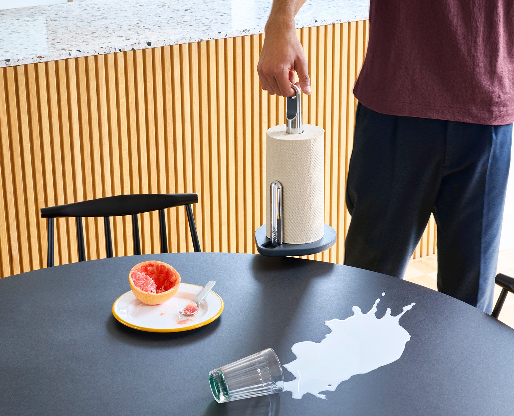 Joseph Joseph Hold Küchenrollenhalter wird an seiner Fingerschlaufe zu verschütteter Milch auf einem Tisch getragen